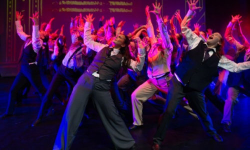 Group of dancers stood in a formation with their hands in the air wearing suits 